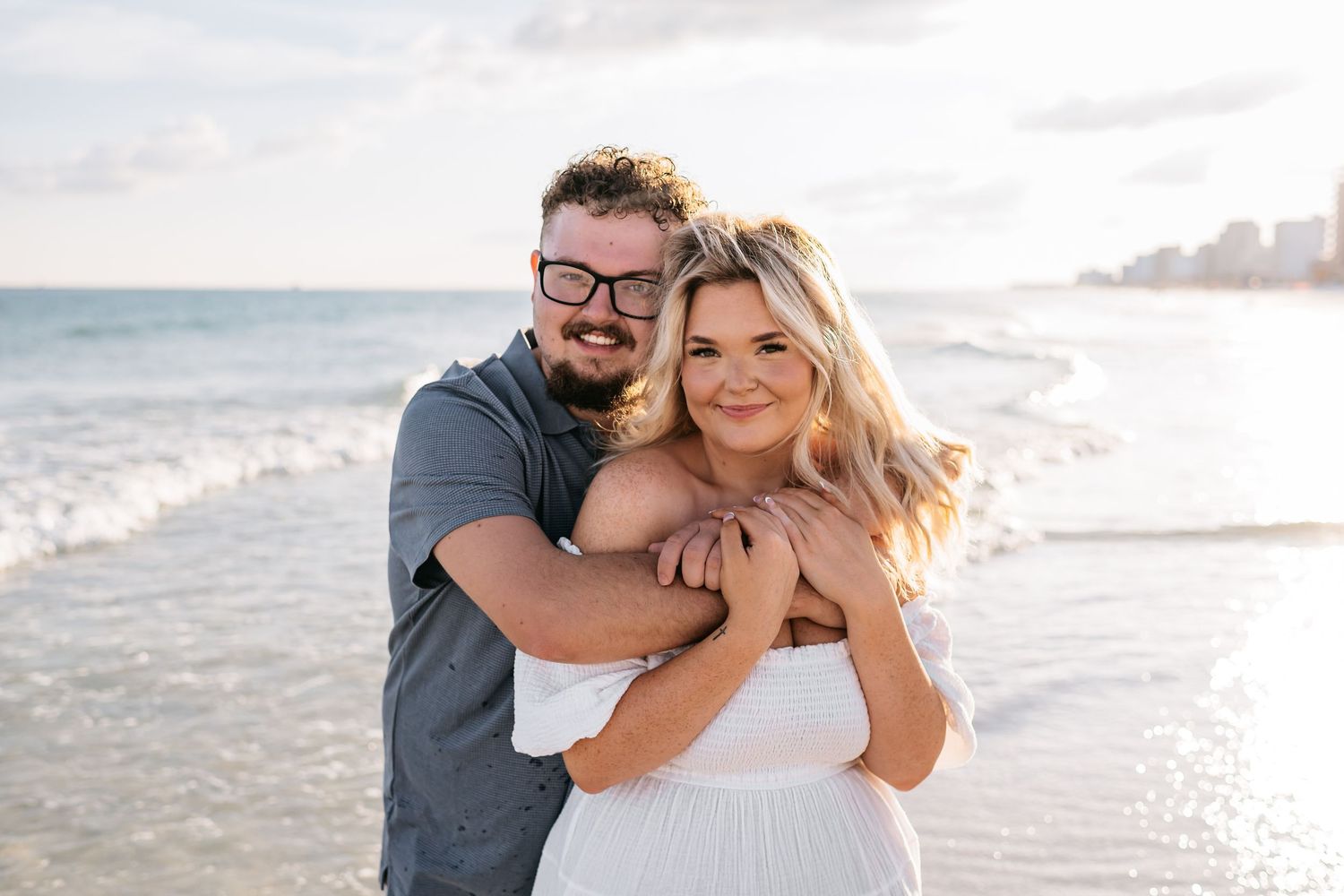Happy couple embracing and laughing together on a sunny beach with coastal scenery.