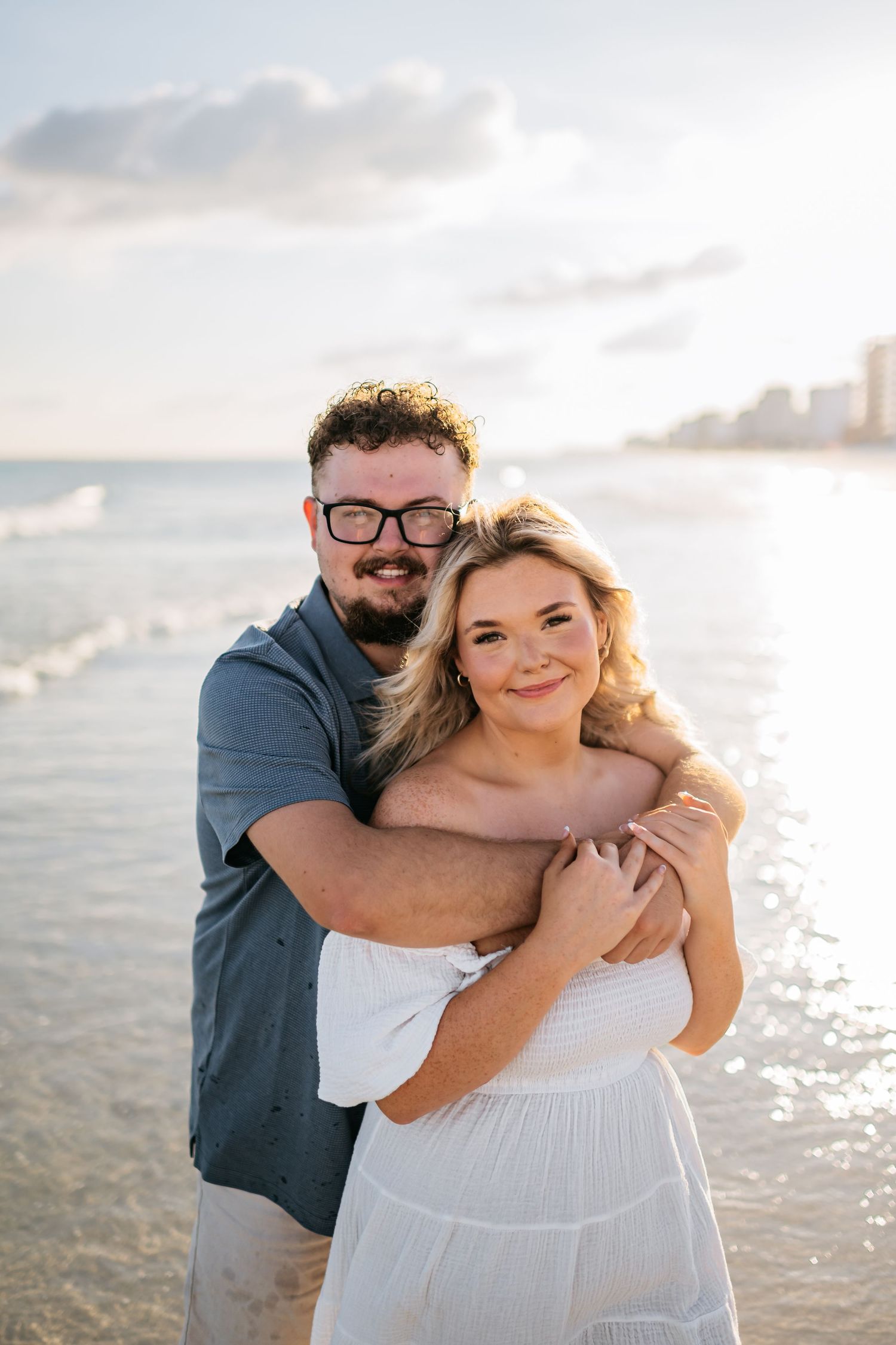 Couple shares intimate moment on beach at sunset with coastal buildings visible in distance.