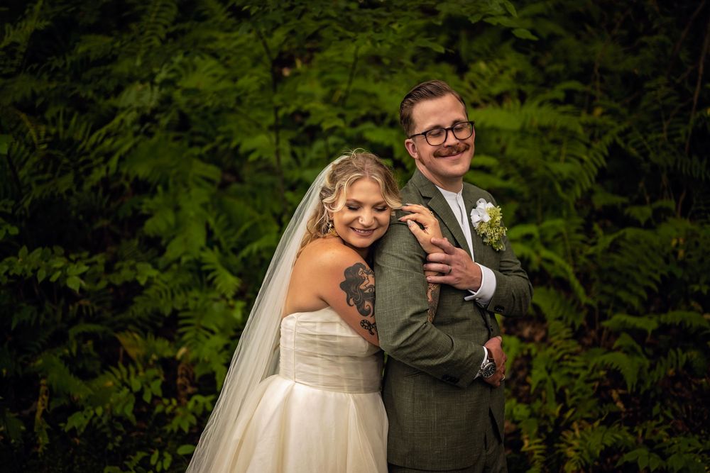 Bride and groom laughing together in front of lush green ferns on their wedding day.