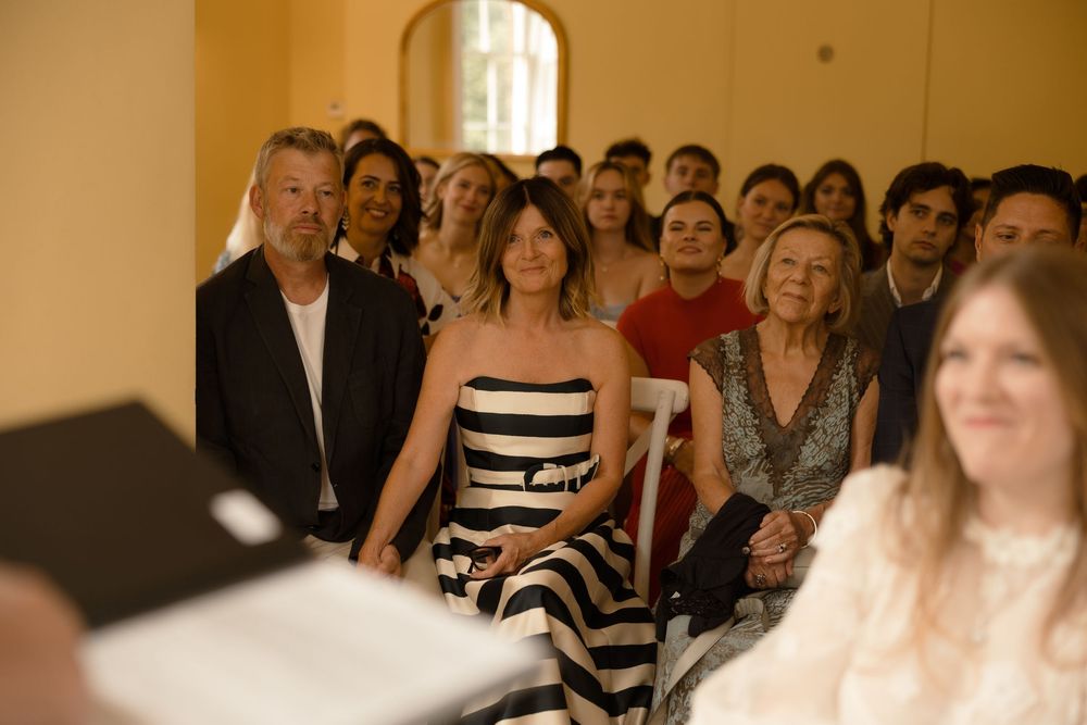 Wedding guests seated in rows at an indoor ceremony venue watch the proceedings with interest.