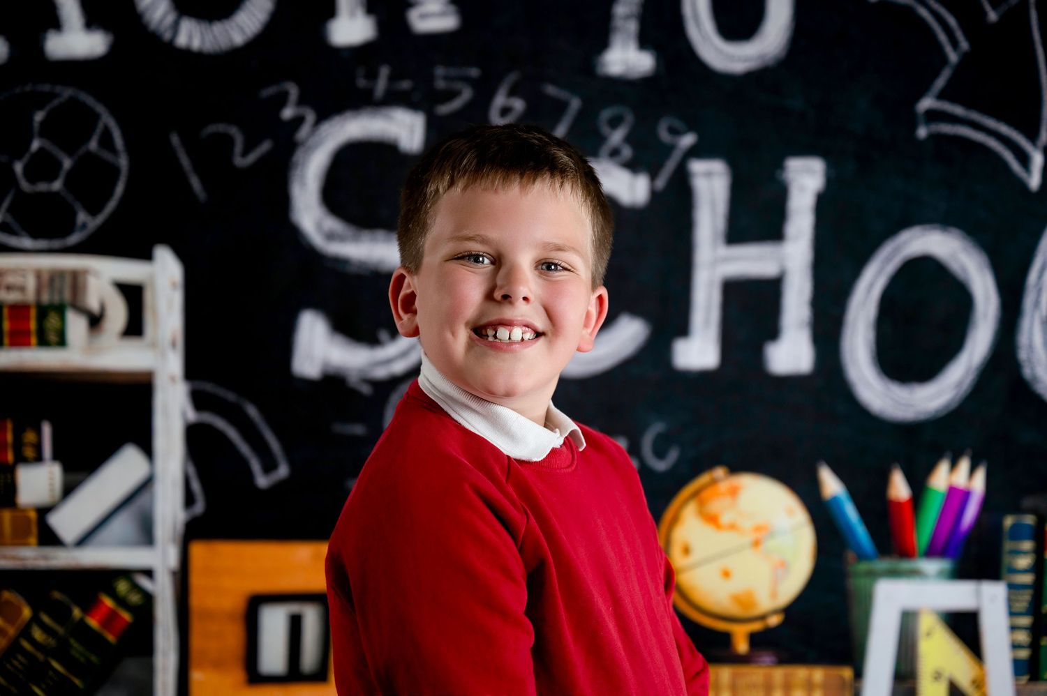 A child in a red sweater stands in front of a classroom chalkboard with educational materials and decorations.