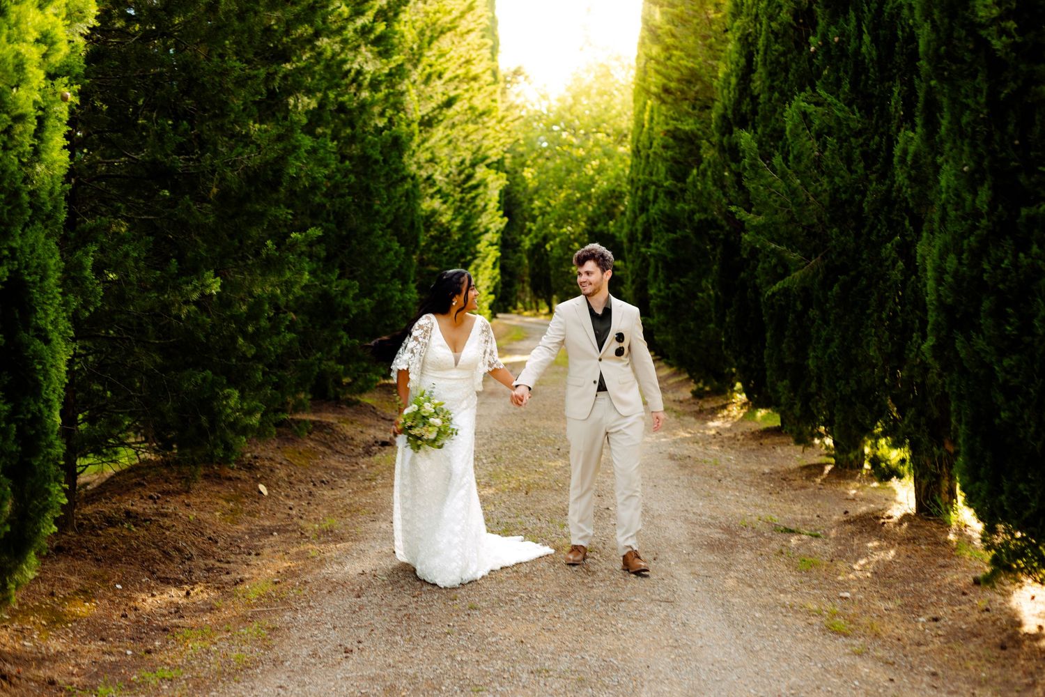 Newlyweds hold hands while walking down tree-lined path at sunset.