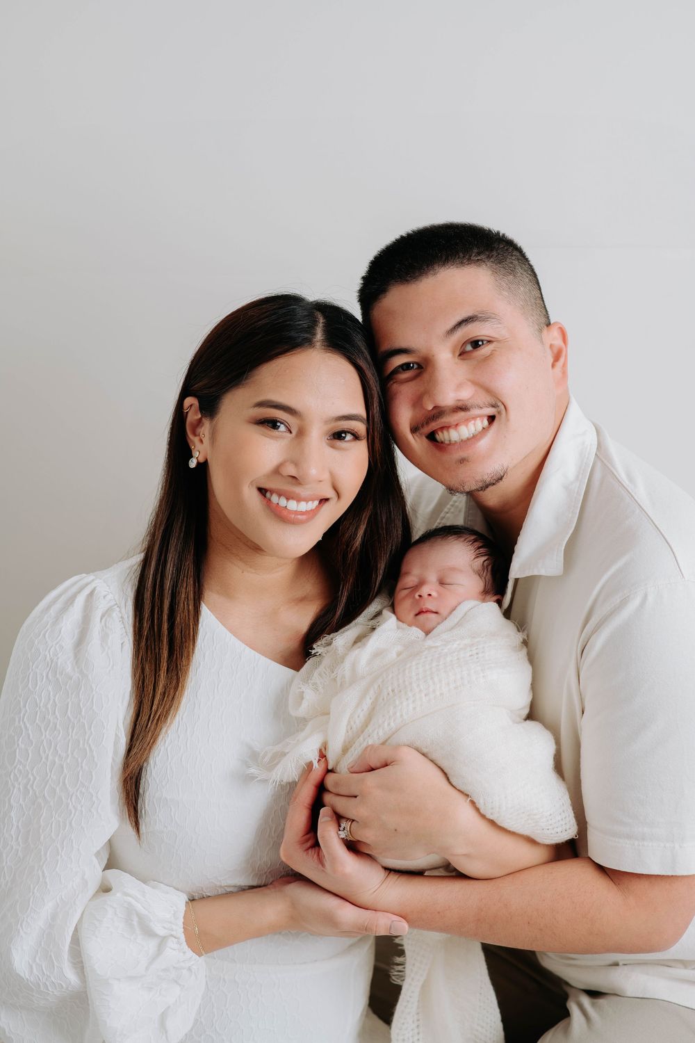 Family wearing white outfits poses together with newborn baby in minimalist studio photoshoot.