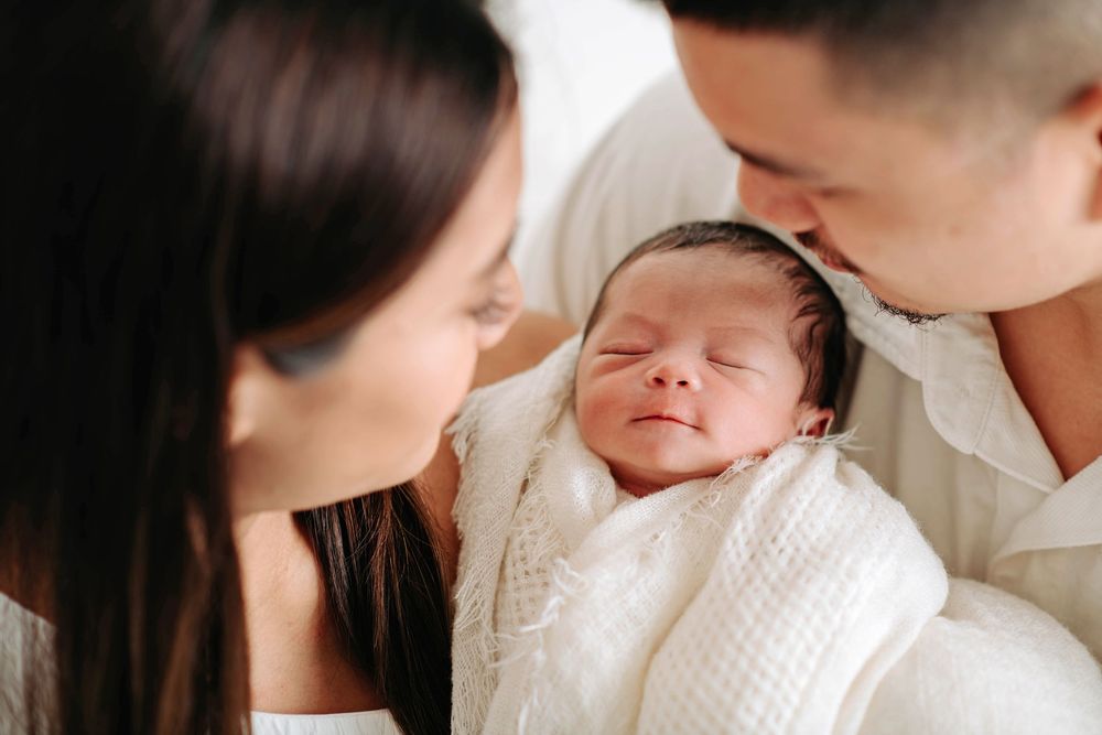 Parents in white clothes looking lovingly at sleeping newborn baby against light background.