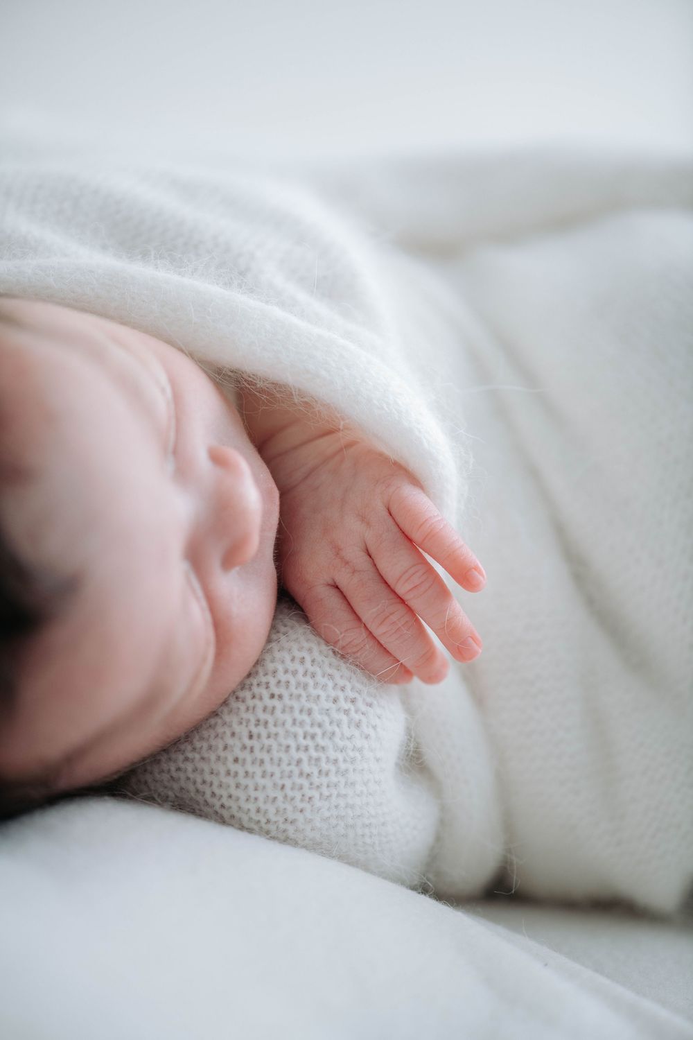 A tiny baby's hand reaches out from soft white blankets in a tender close-up shot.