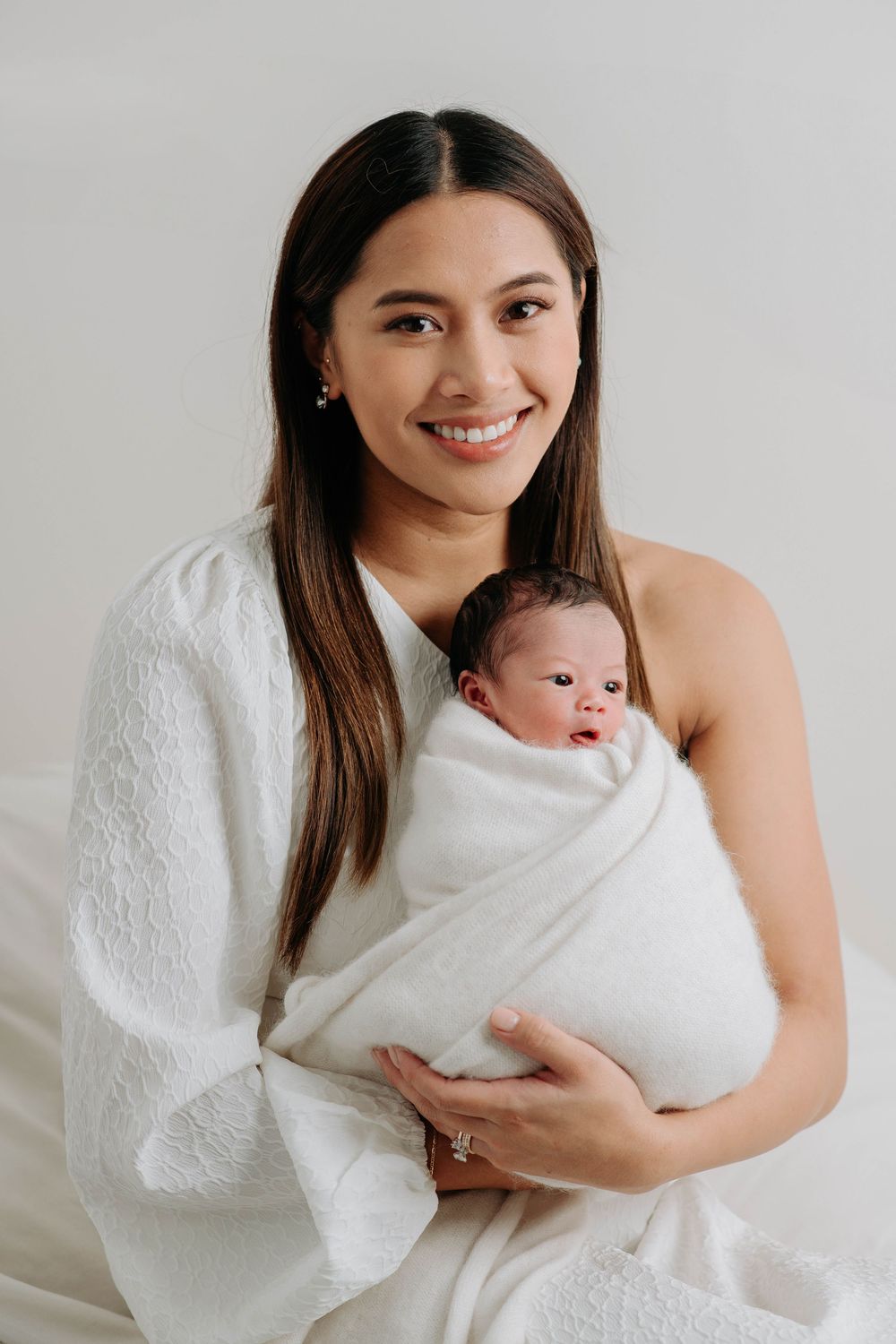 Mother wearing white holds sleeping newborn wrapped in soft towel against neutral background.