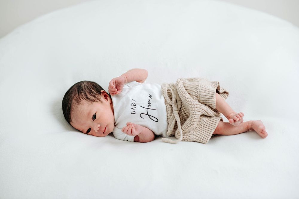 Newborn baby lying on white blanket wearing tiny white onesie and beige cardigan.