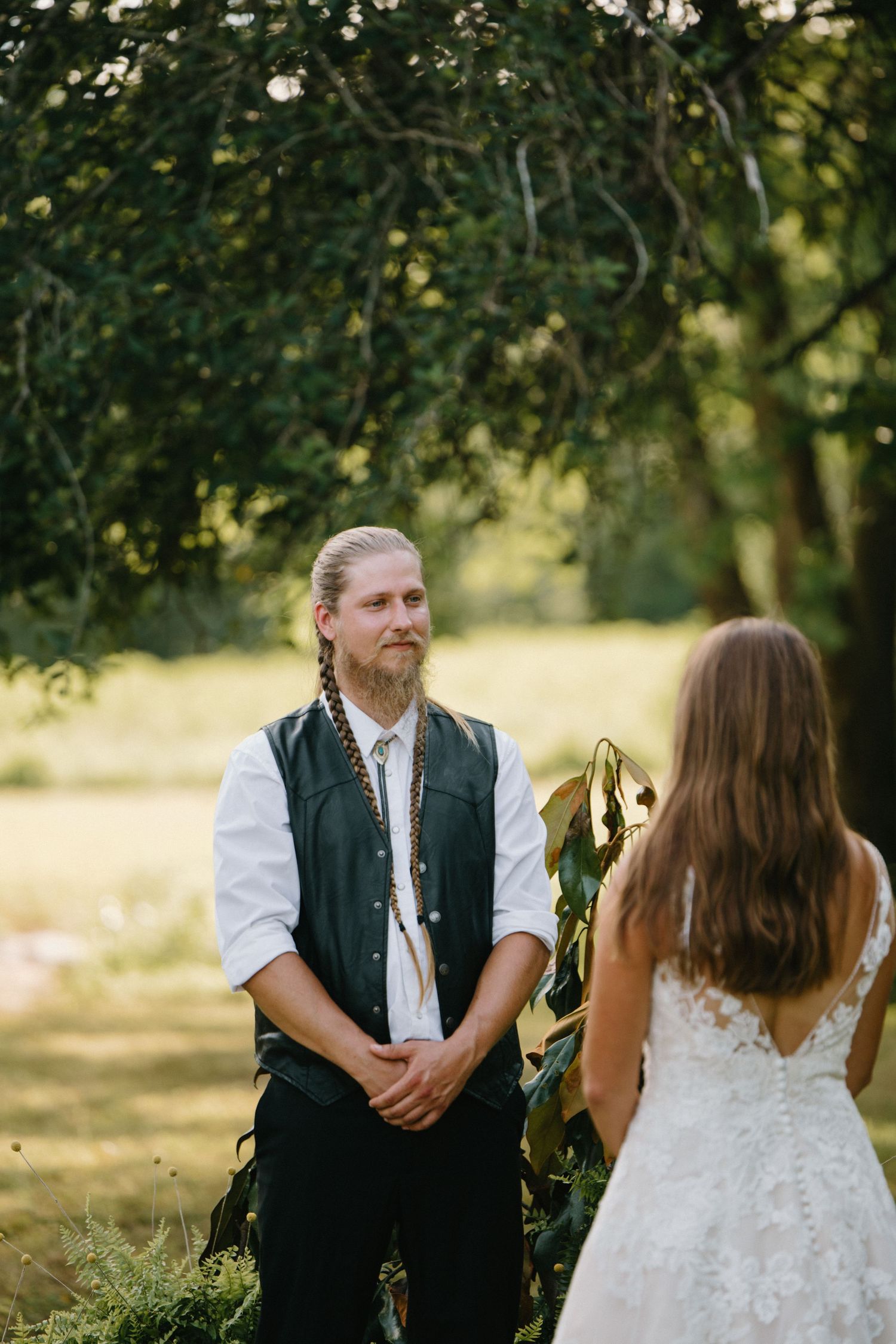 A couple shares a tender moment outdoors under sunlit trees at a rustic wedding celebration.
