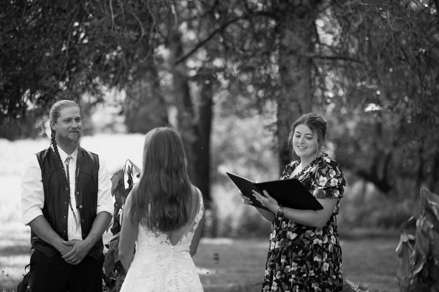 A couple exchanges vows during an intimate outdoor wedding ceremony while an officiant in floral dress reads from a binder.