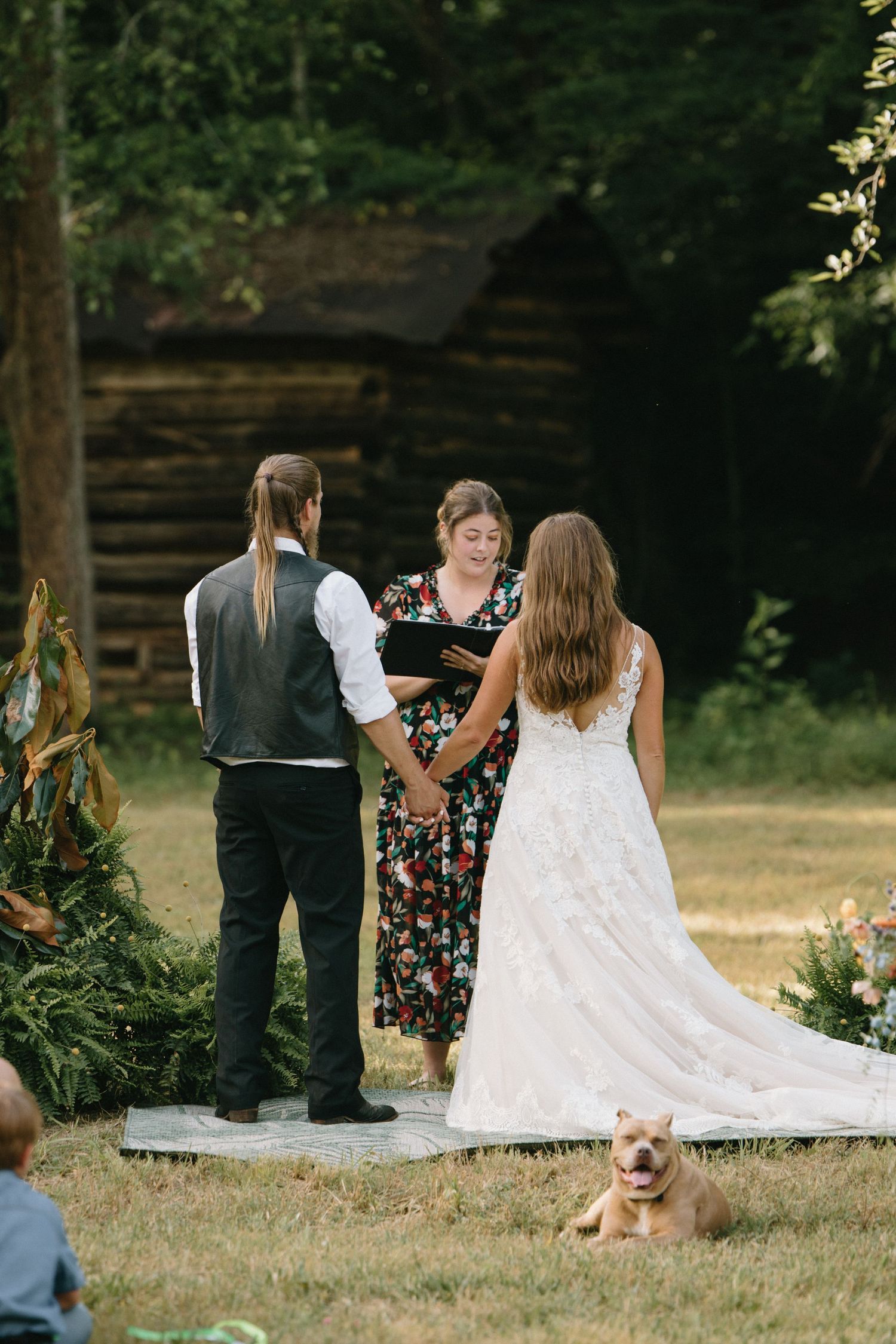 An intimate outdoor wedding ceremony in front of a rustic log cabin with a small dog sitting in the foreground.