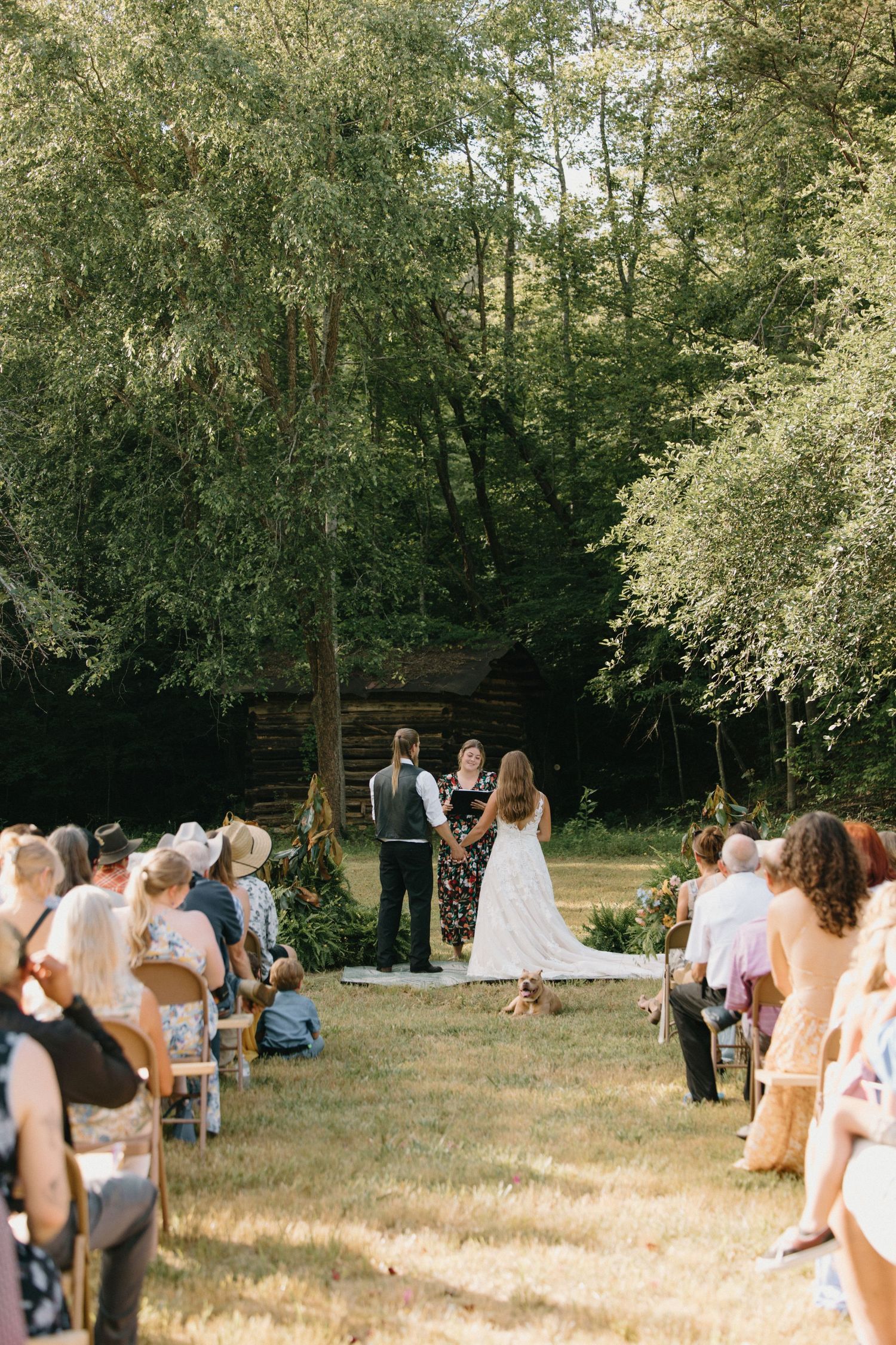 Outdoor wedding ceremony in a wooded area with guests seated on rustic wooden benches under sun-dappled trees.
