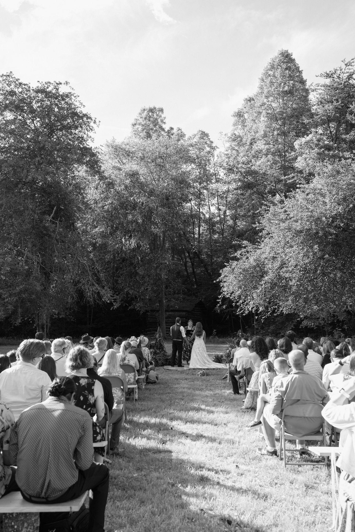 A black and white panoramic view of an outdoor wedding ceremony with guests seated on a lawn surrounded by trees.