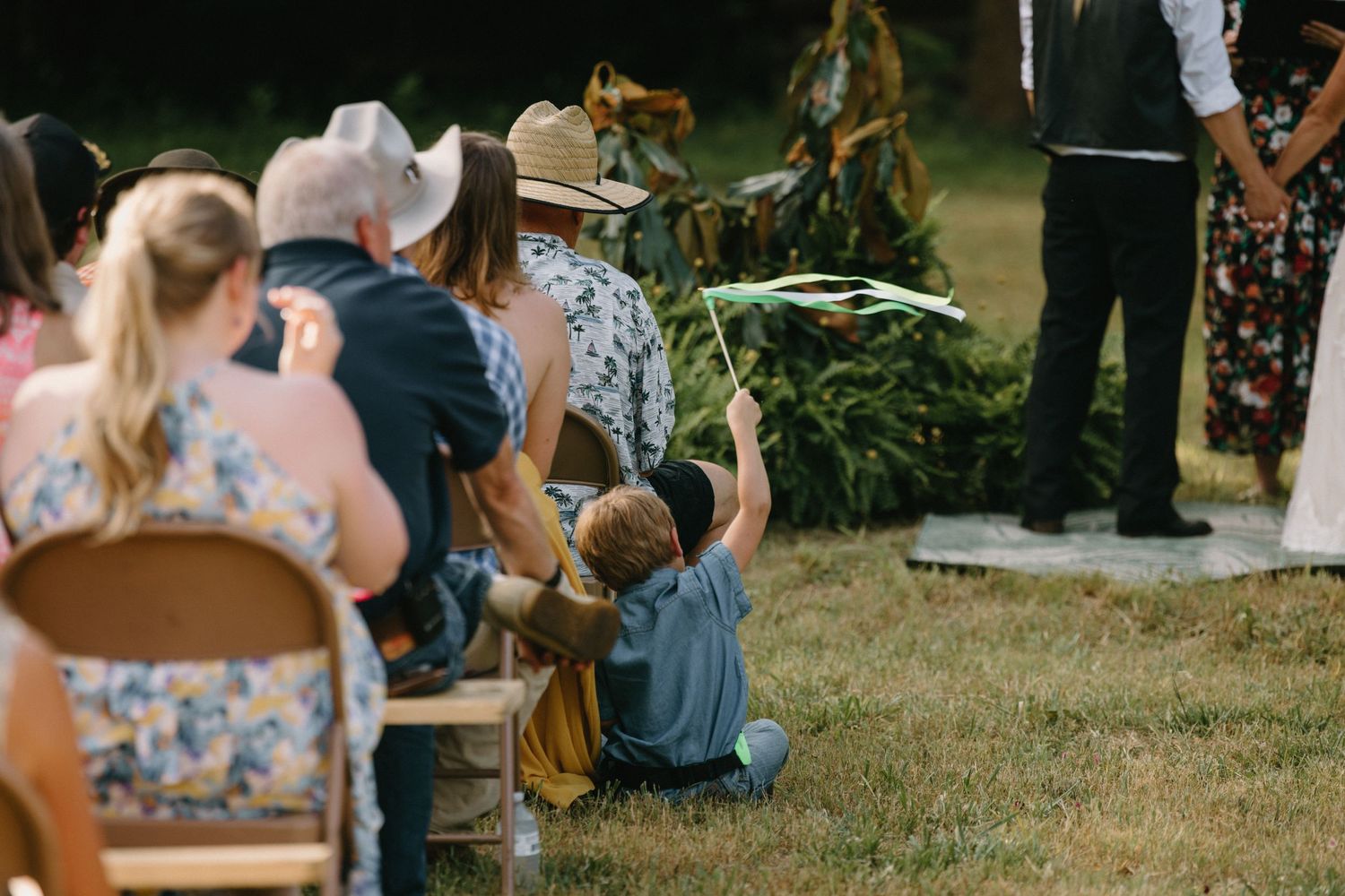 Wedding guests sit in wooden folding chairs on a grassy lawn during an outdoor ceremony.