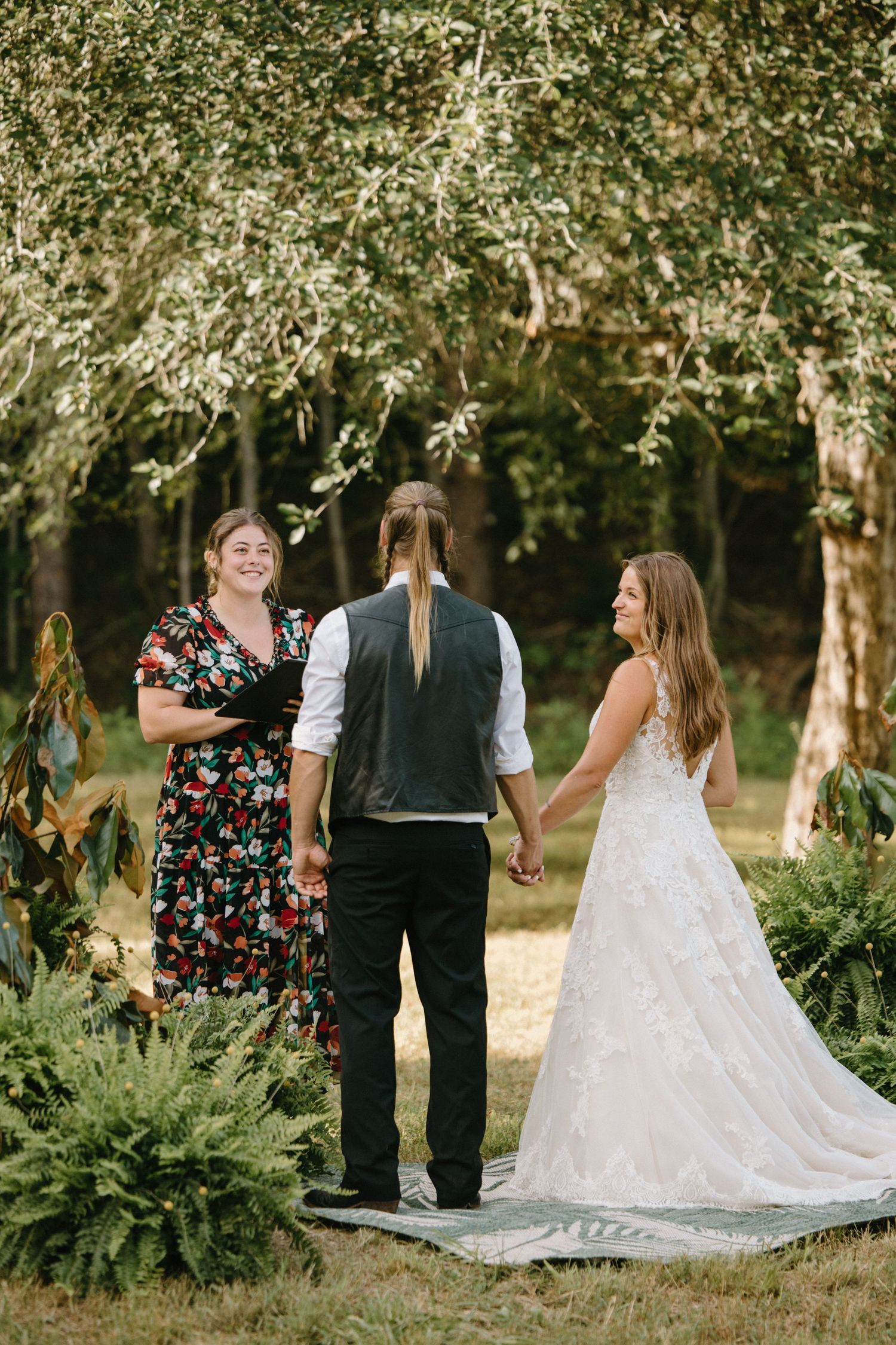 Outdoor wedding ceremony under flowering trees with bride in white lace gown and officiant in floral dress.