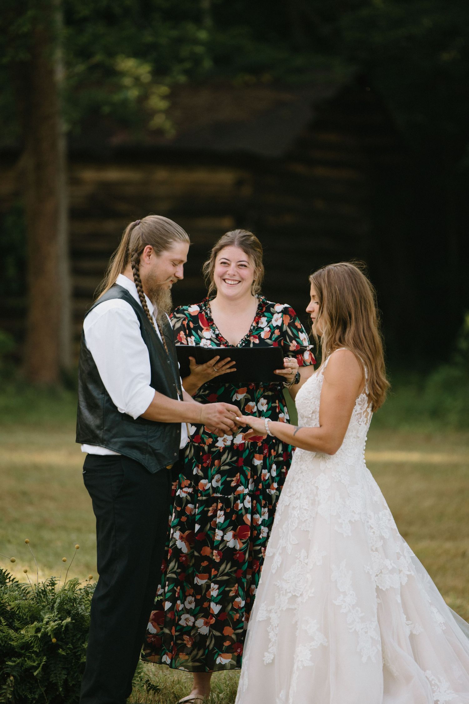 A wedding ceremony takes place outdoors in a wooded setting with an officiant and couple exchanging vows.