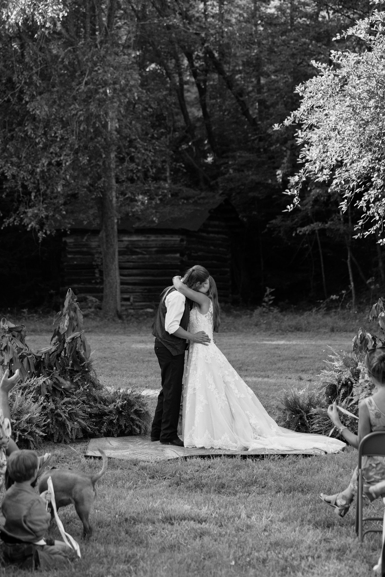 A black and white photograph shows a romantic moment between a couple in wedding attire standing in a garden setting.