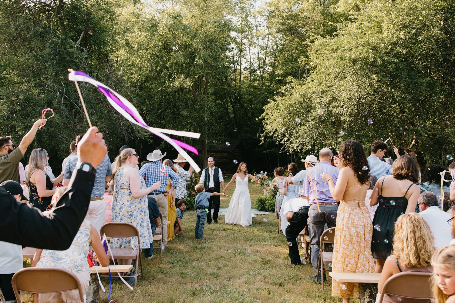 Wedding guests create an archway with raised arms as couple exits outdoor summer ceremony in wooded setting.