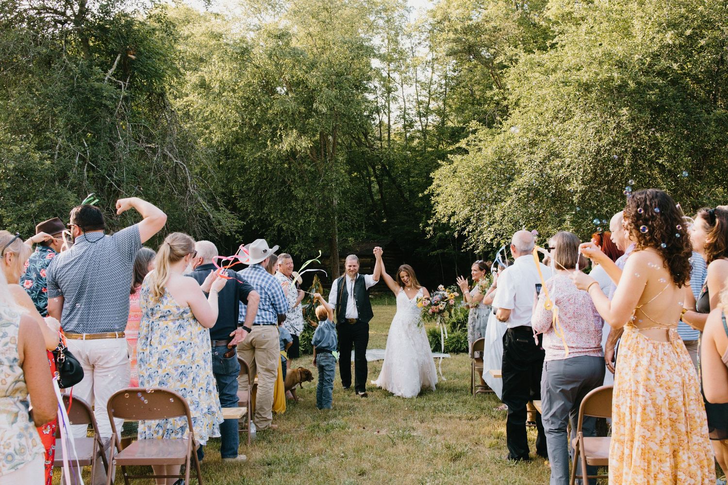 Wedding guests gather for an outdoor ceremony in a picturesque wooded setting with warm natural lighting.