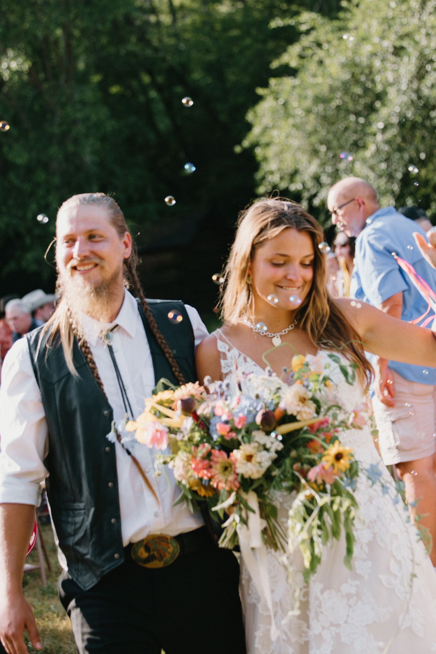 Wedding party celebrating outdoors in bright sunlight with bubbles floating in the air and colorful bouquets.