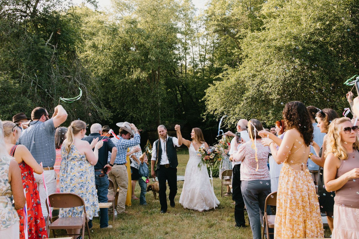 A wedding celebration takes place outdoors in a garden setting with guests mingling under sunlit trees.