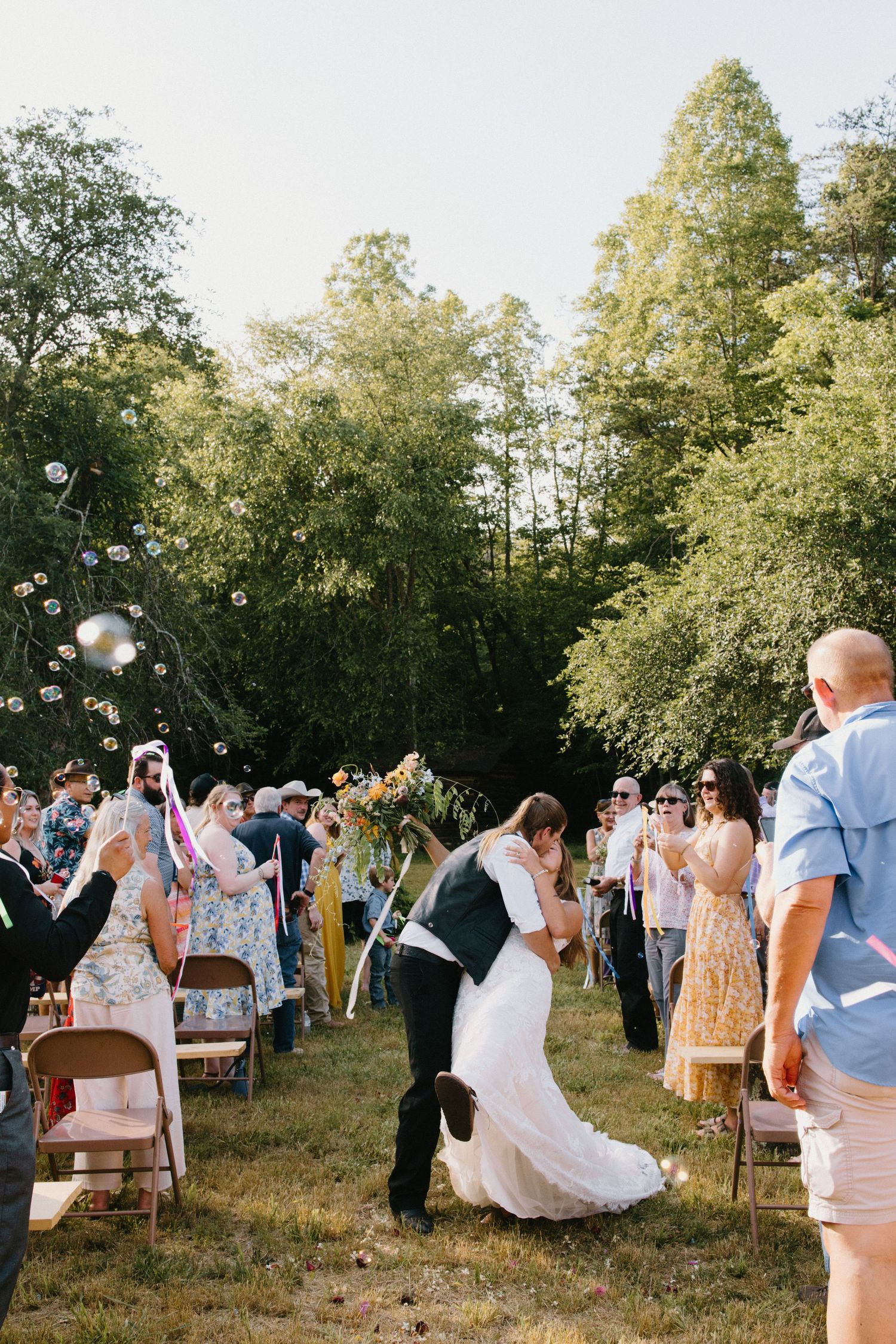 Wedding guests blow soap bubbles during an outdoor summer celebration in a sunlit grove.