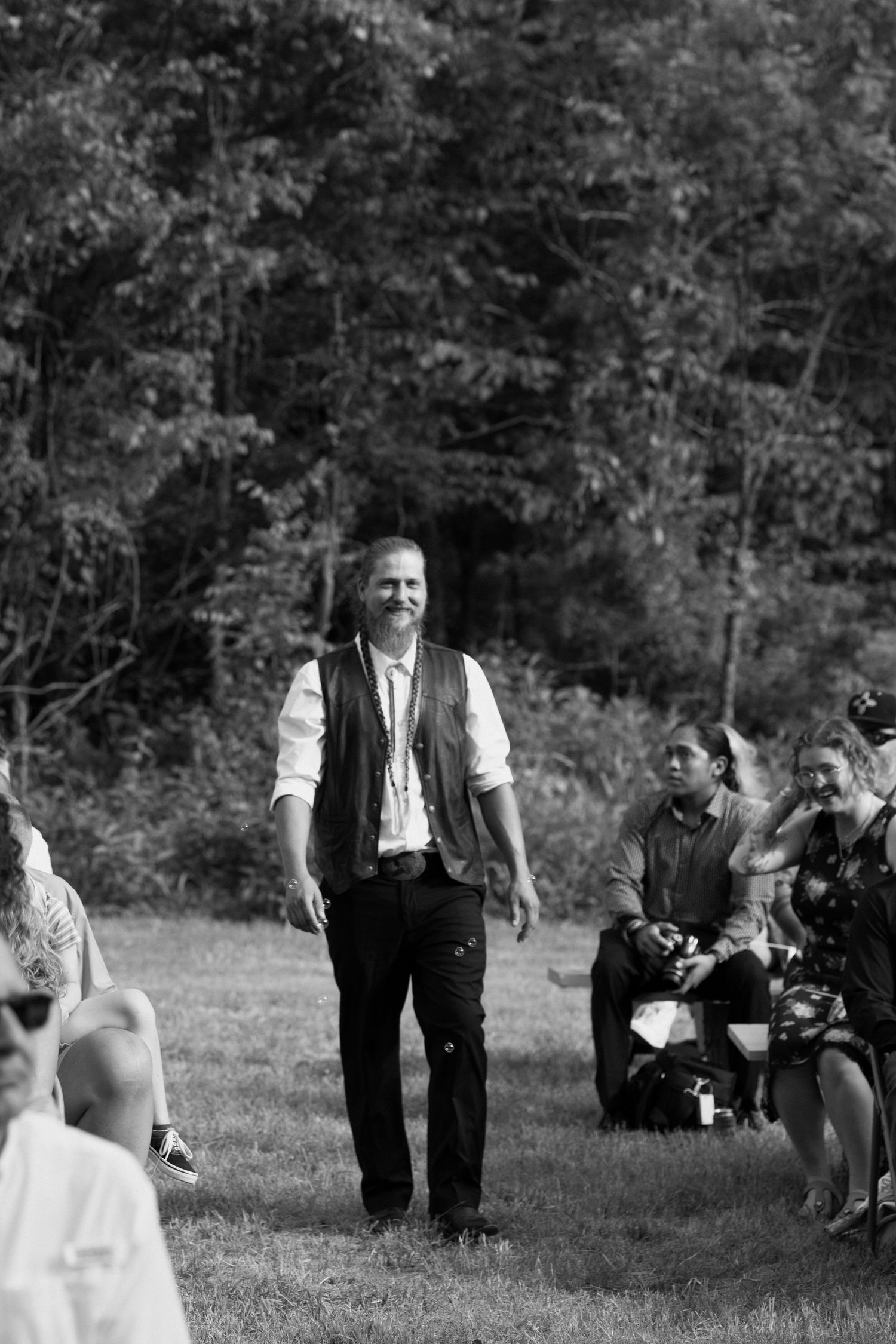 A black and white photo shows a person in a vest and dress shirt standing outdoors while others sit on the grass.