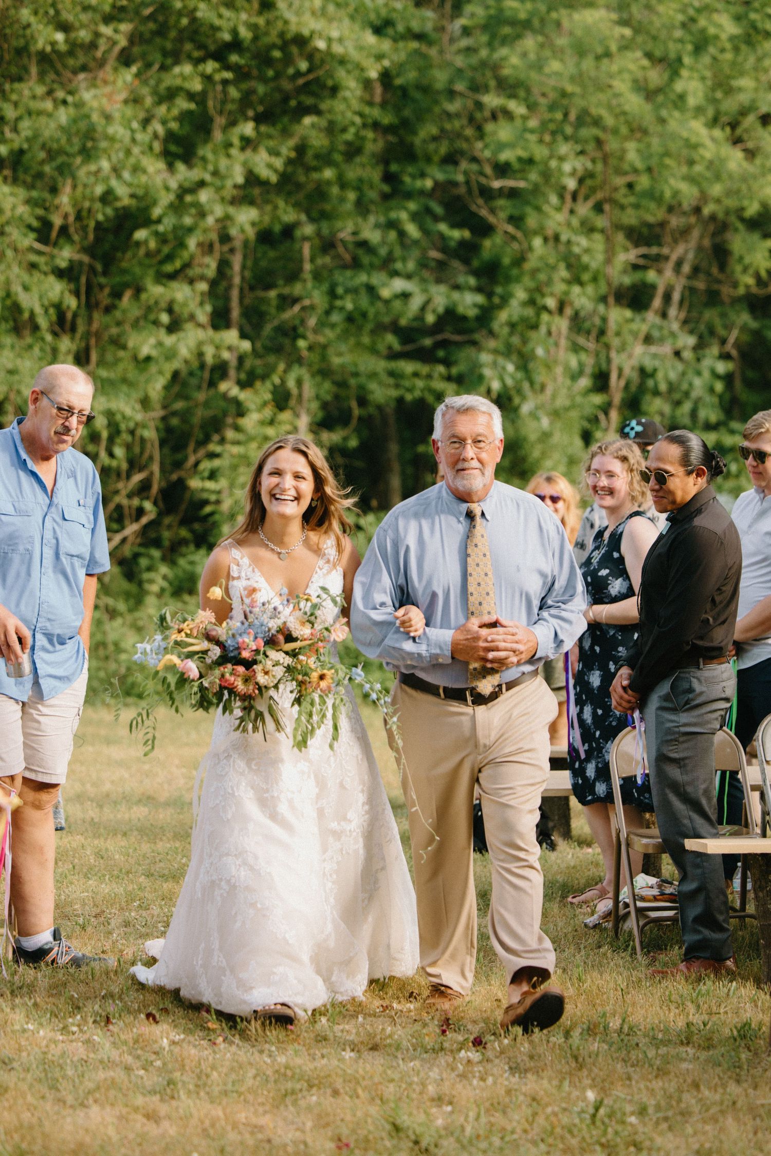 A joyful group moves down a grassy path at an outdoor wedding celebration with attendees in light summer attire.
