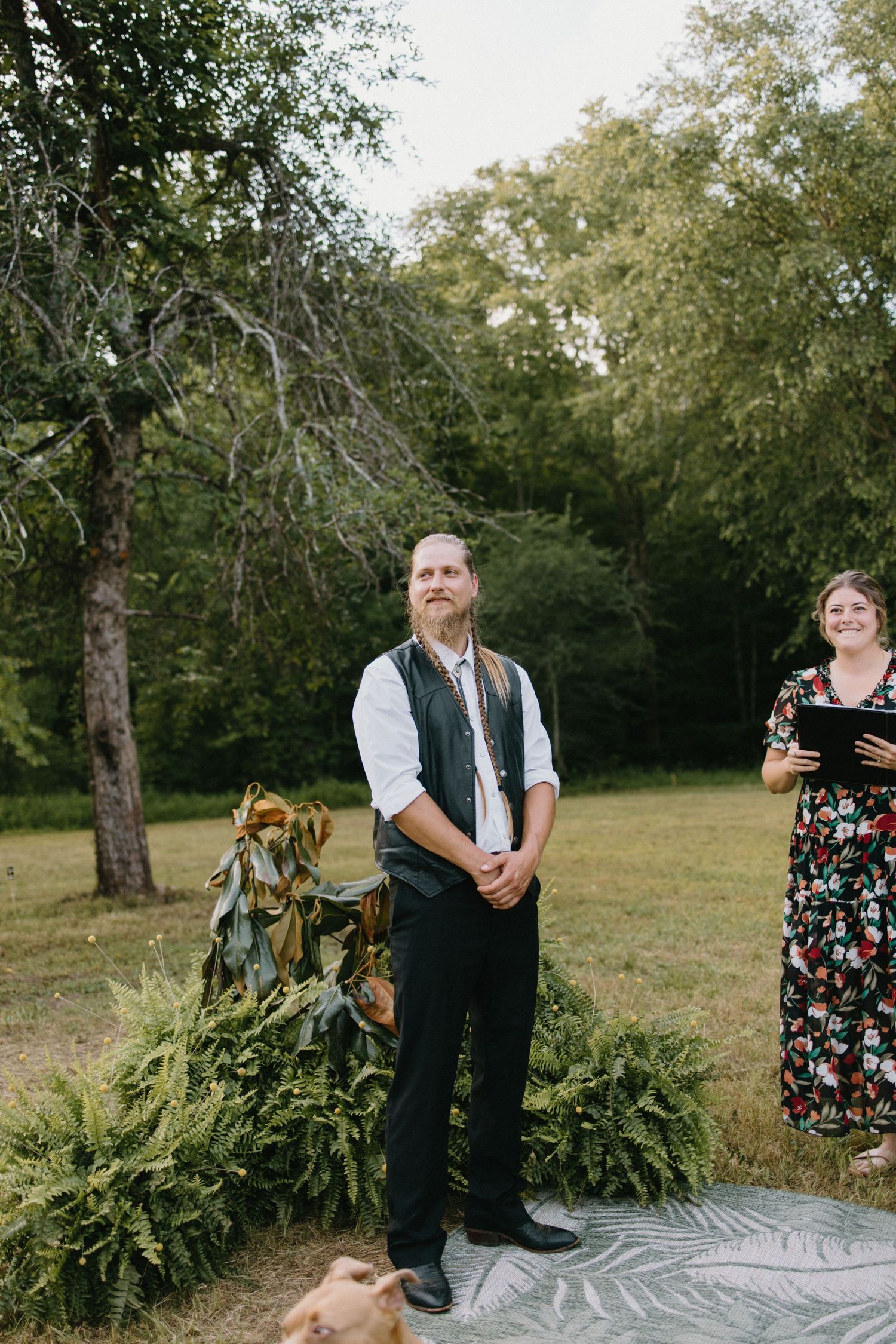 A couple stands together on a picturesque patterned blanket in a grassy meadow surrounded by trees.