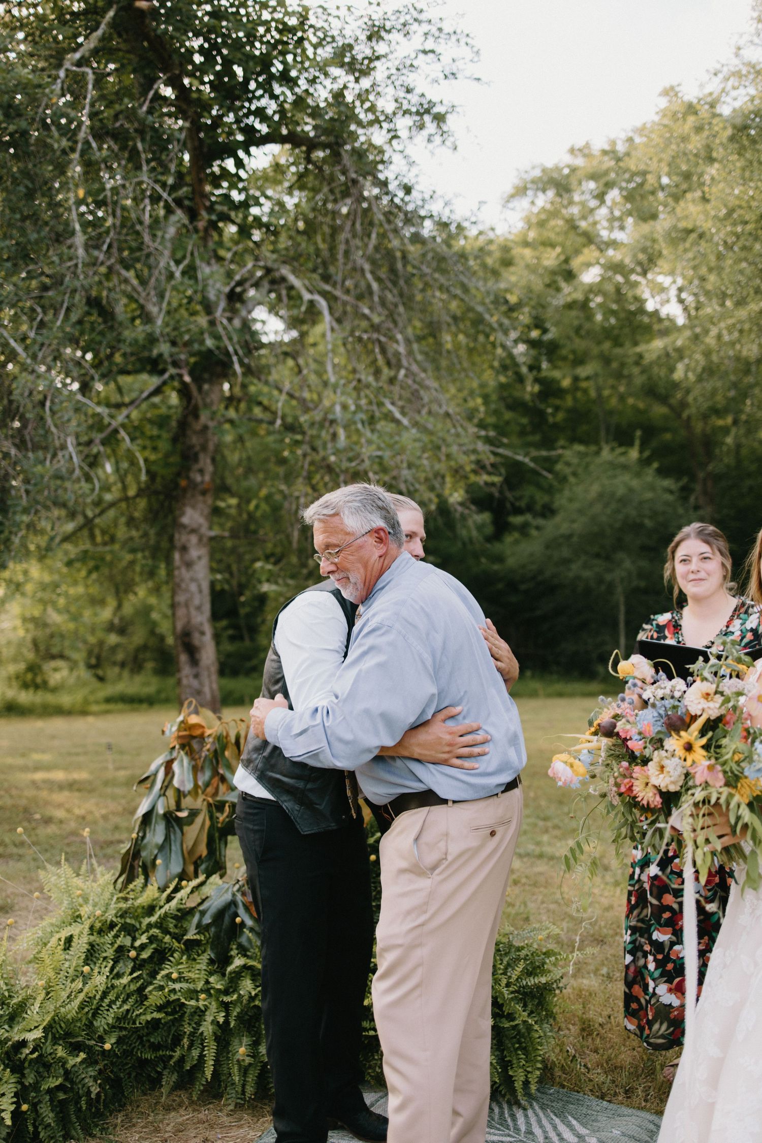 Two people embrace warmly at an outdoor wedding ceremony with floral decorations amid trees in natural sunlight.