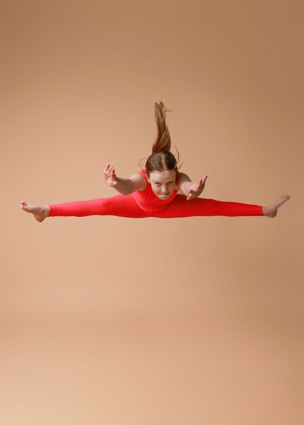 Dancer in bright red leotard performs a graceful split jump against a neutral beige background.