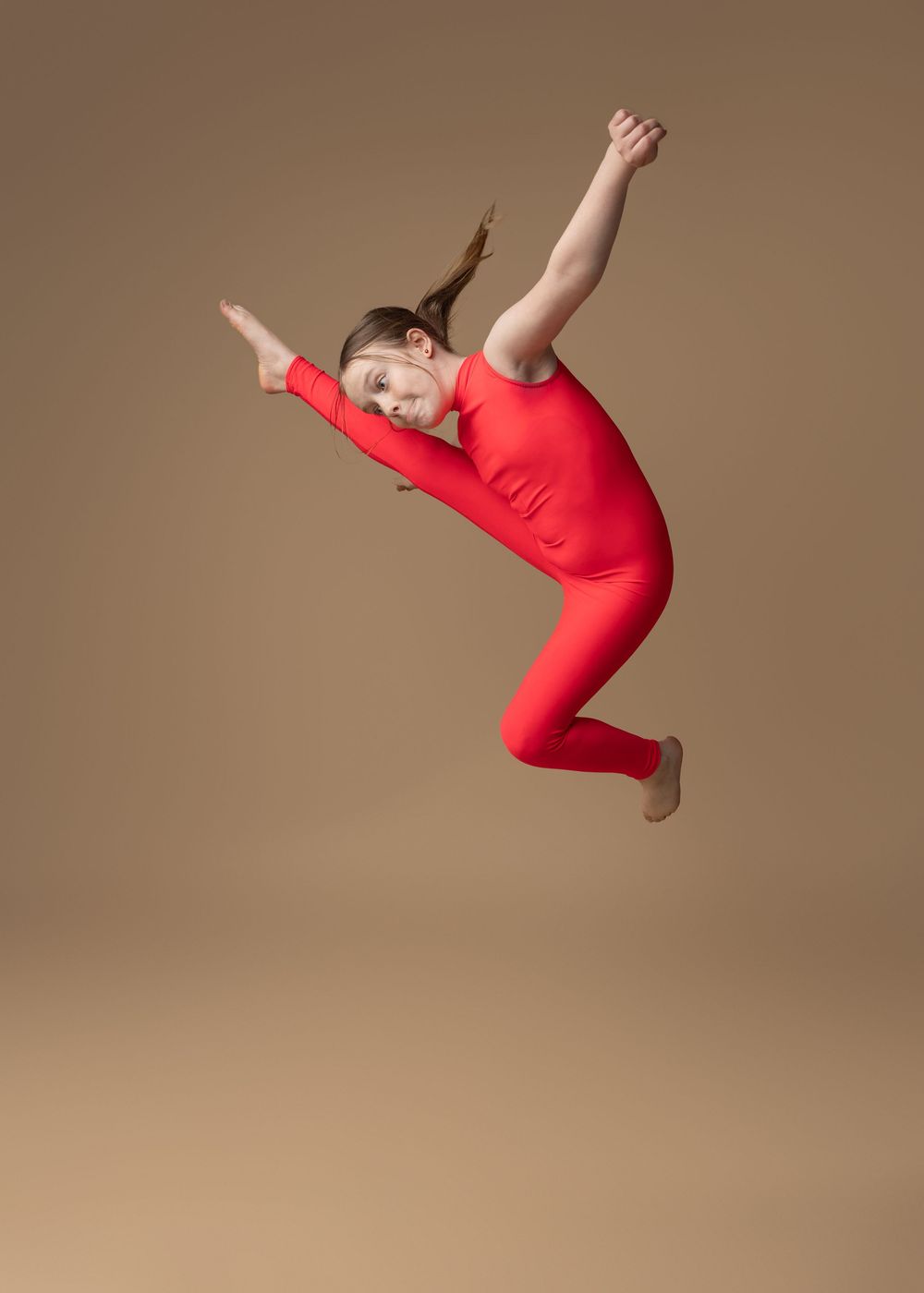 Dancer in bright red unitard performs graceful aerial jump pose against neutral beige background.