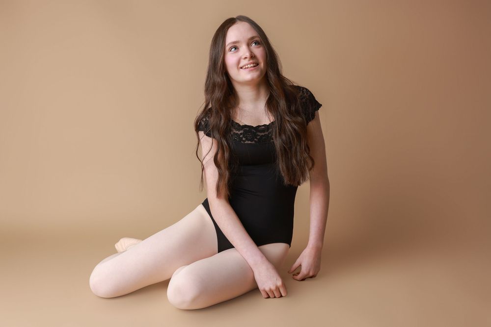 Ballet dancer in black leotard sitting gracefully on studio floor with light brown backdrop.