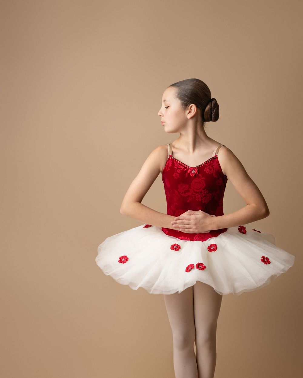 Ballet dancer in a red velvet bodice and white tulle tutu with red floral accents poses elegantly against beige background.