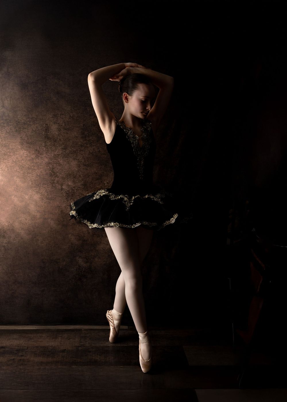 A ballet dancer in a black tutu poses gracefully en pointe against a dark dramatic background with moody lighting.