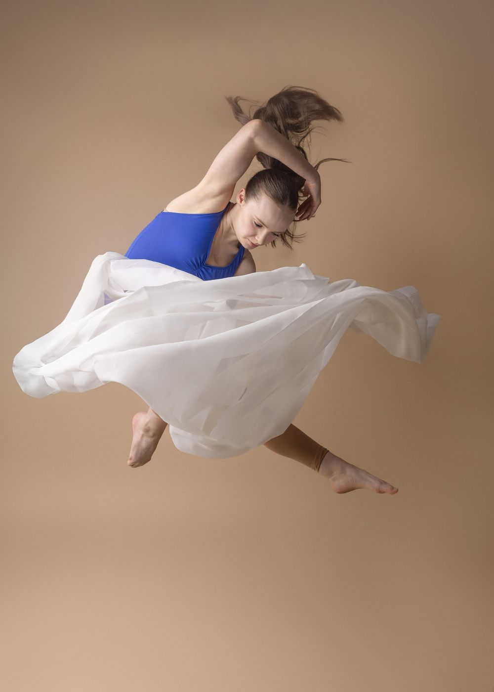 A dancer in a blue leotard and flowing white skirt leaps gracefully against a neutral beige backdrop during a performance.