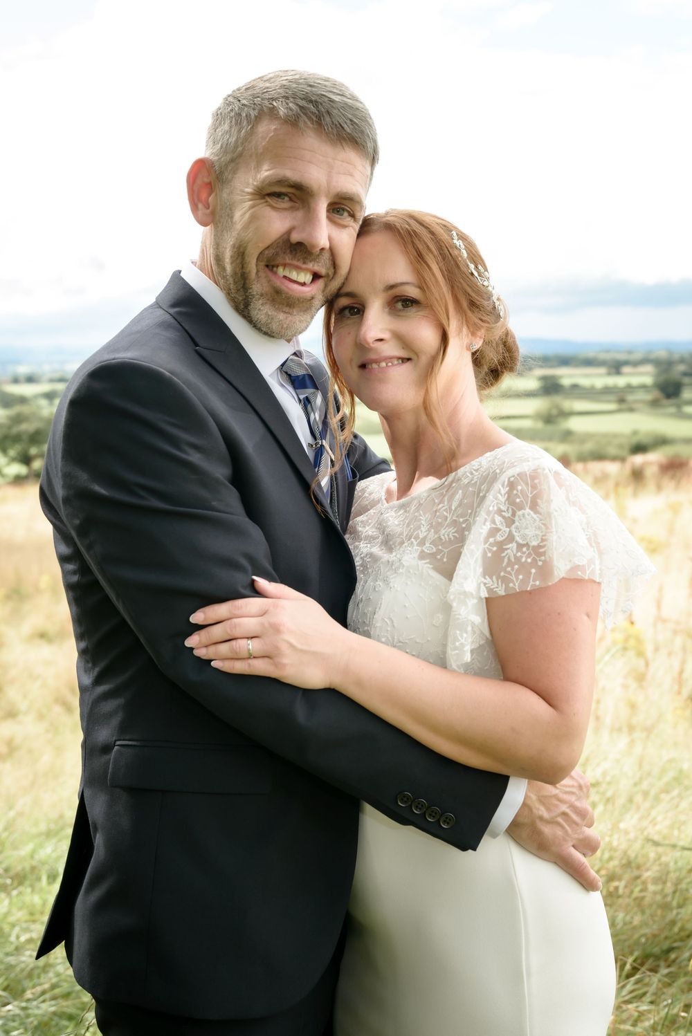 Happy wedding couple embraces in grassy field with rolling hills and countryside landscape in background.