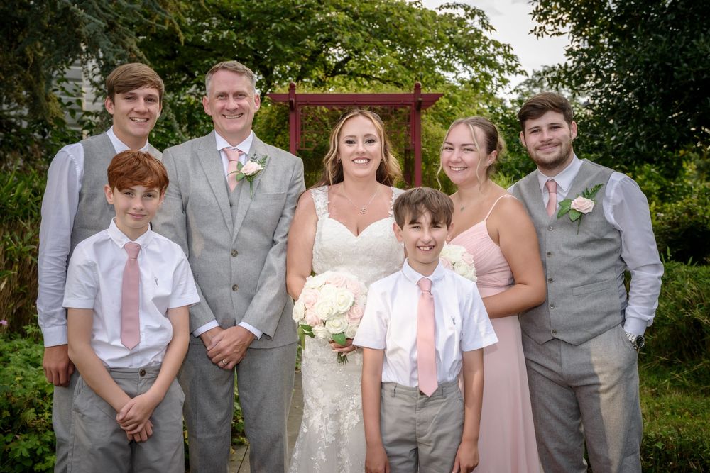 Wedding party in grey suits and pink dresses poses in front of garden arbor with bride holding white bouquet.