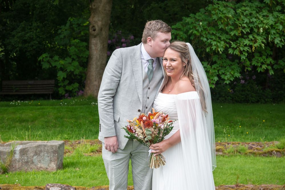 A couple share a tender moment on their wedding day in a lush garden setting, the bride holding a vibrant orange bouquet.