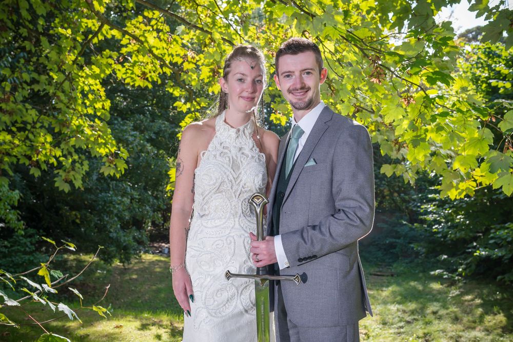 A bride and groom pose together outdoors under sunlit green leaves in a natural garden setting.