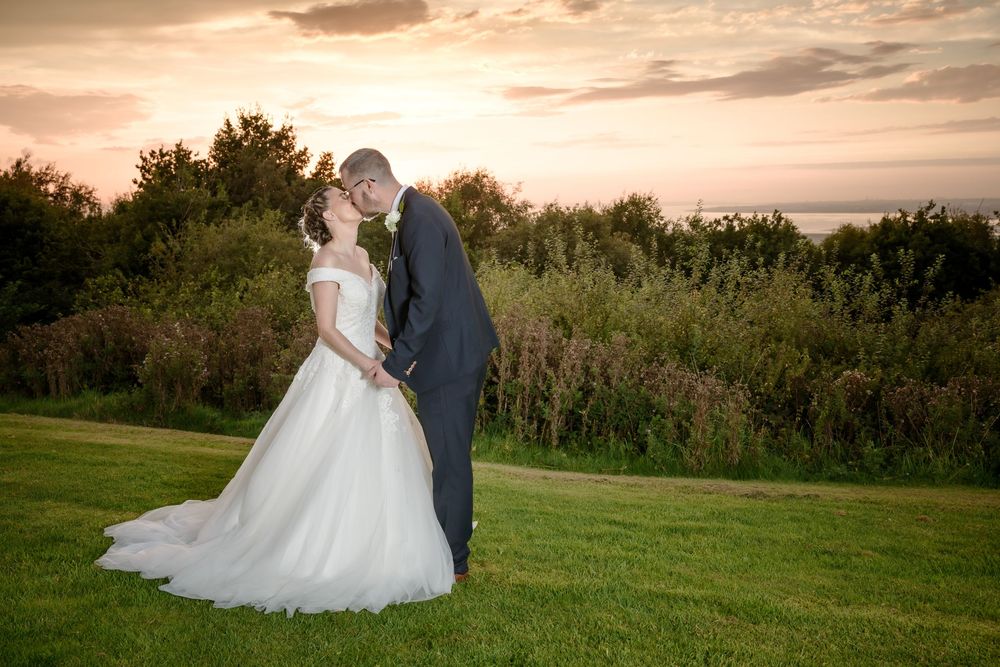 A romantic sunset wedding portrait on a grassy hilltop with a couple embracing against a golden sky and green foliage.