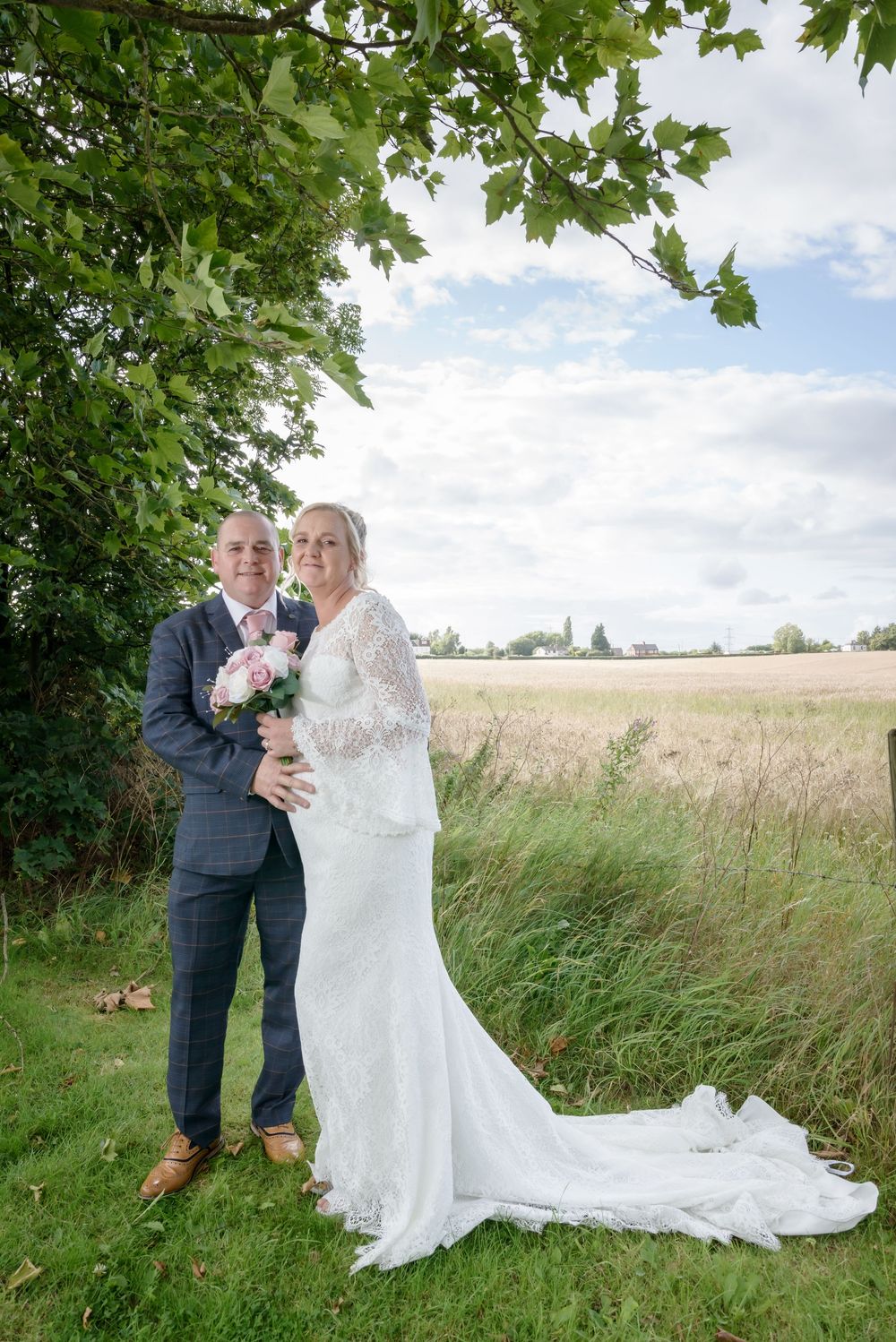 A bride and groom pose together in a beautiful outdoor setting with green trees and a wheat field in the background.