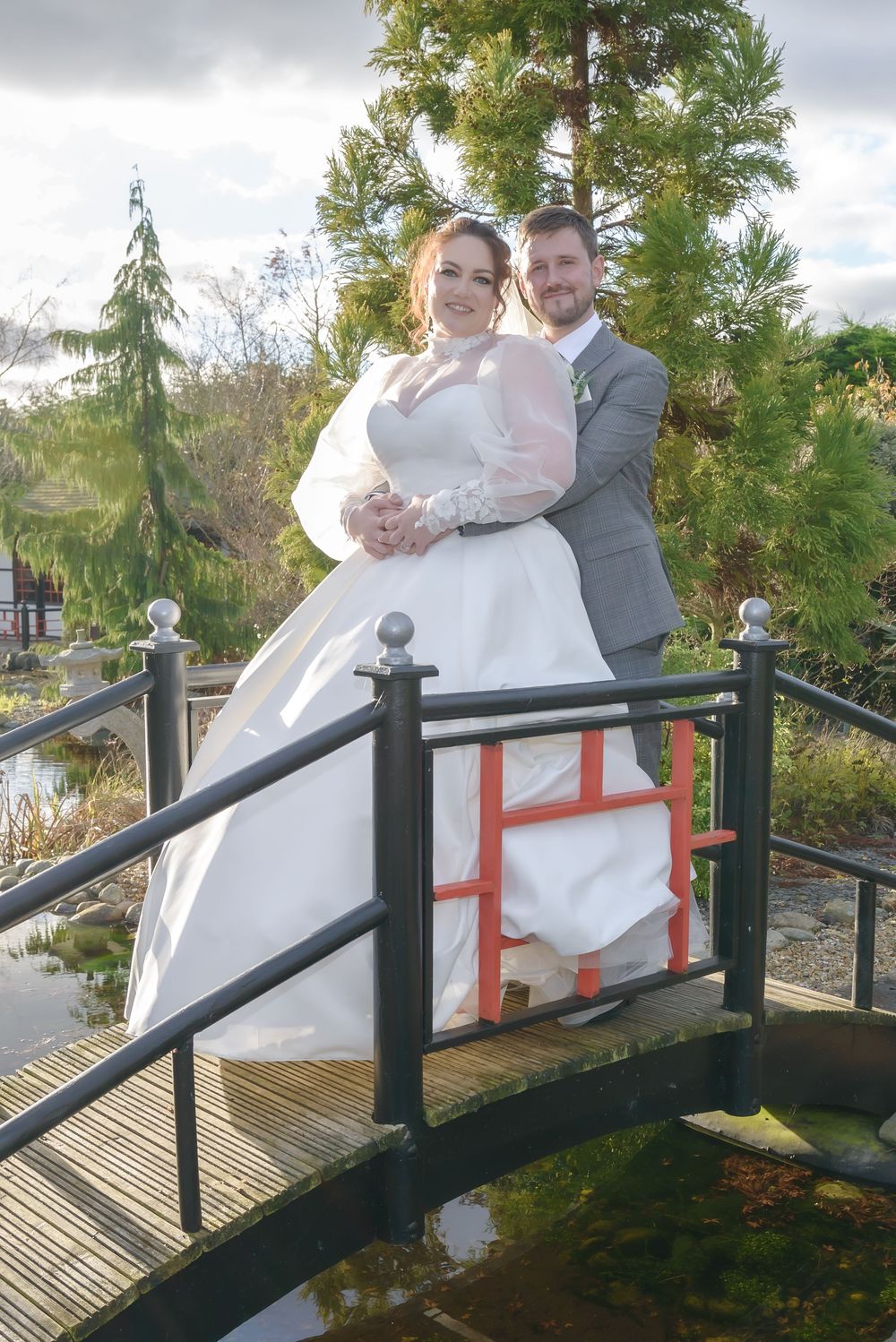 A couple stands on a wooden footbridge in an outdoor garden setting on their wedding day.