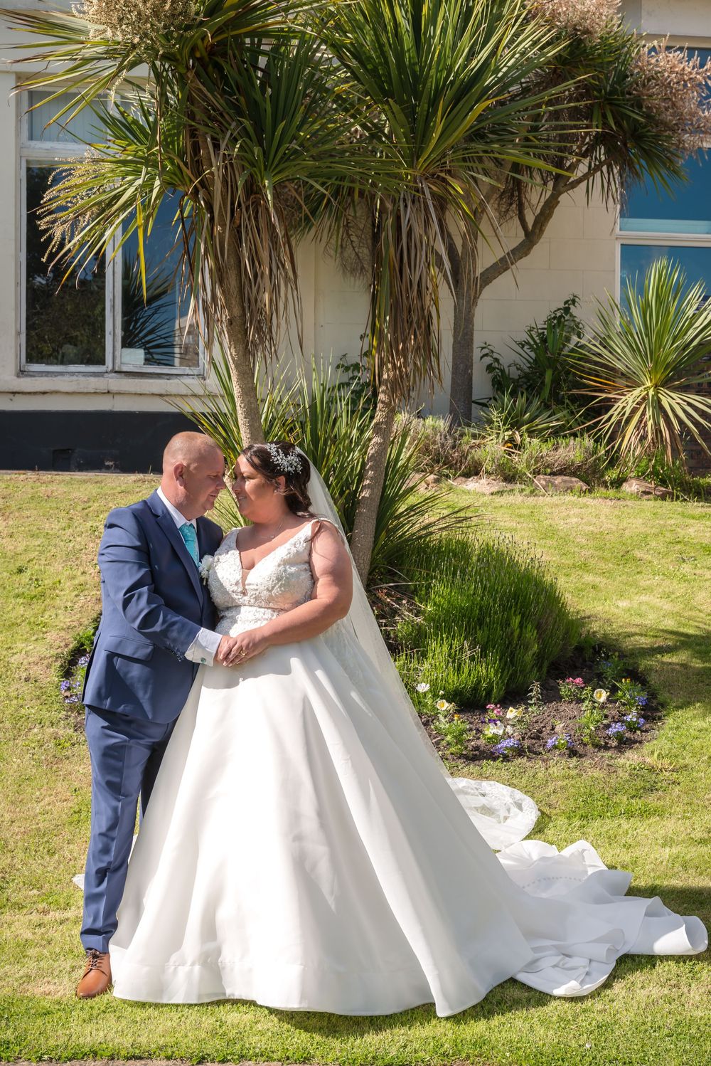 A couple in formal wedding attire embraces on a sunny lawn with palm trees and a building in the background.