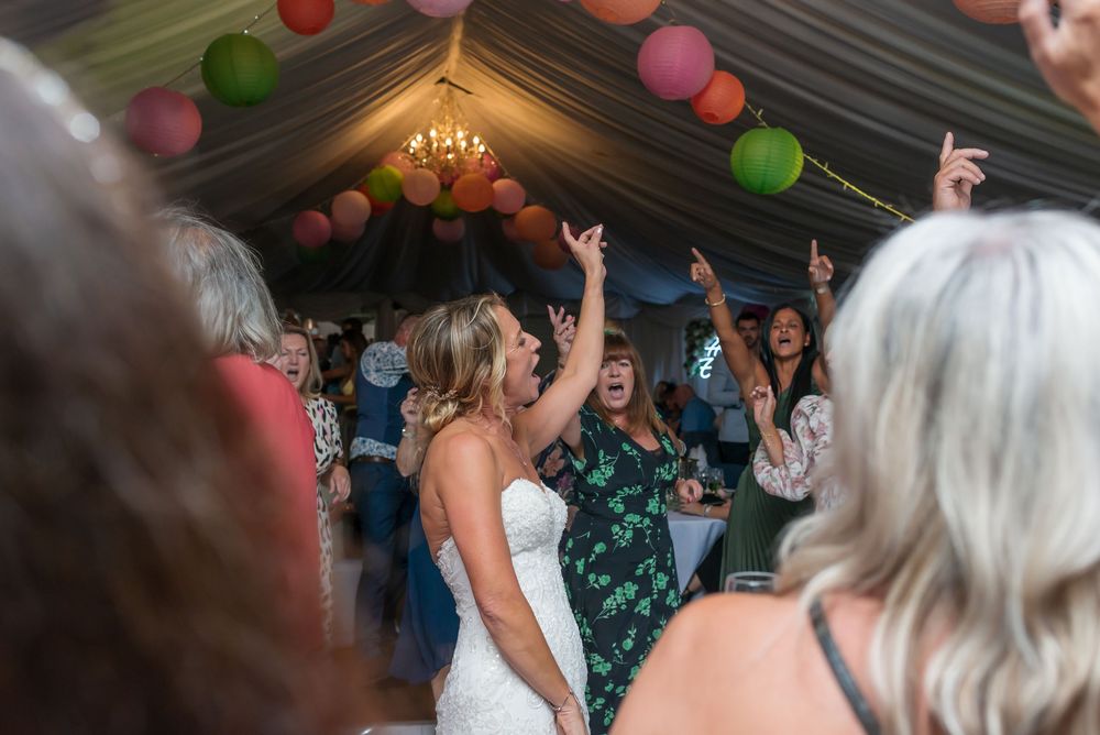 A lively wedding reception celebration under a tent with colorful balloons as guests dance and raise their hands.