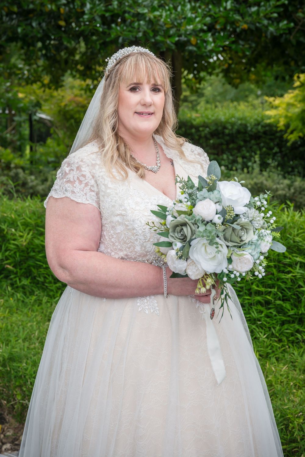 Beautiful wedding portrait featuring a bride in a lace and tulle gown holding an elegant white rose bouquet in a garden setting.