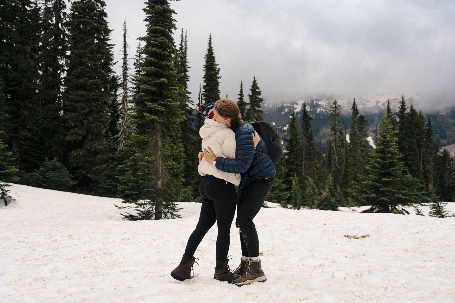 Two people embrace in a snowy winter landscape surrounded by evergreen trees and mountain peaks in the distance.