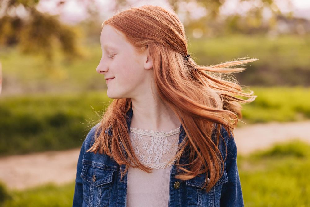 Person with flowing red hair wearing denim jacket stands in sunlit field with wind-swept tresses.