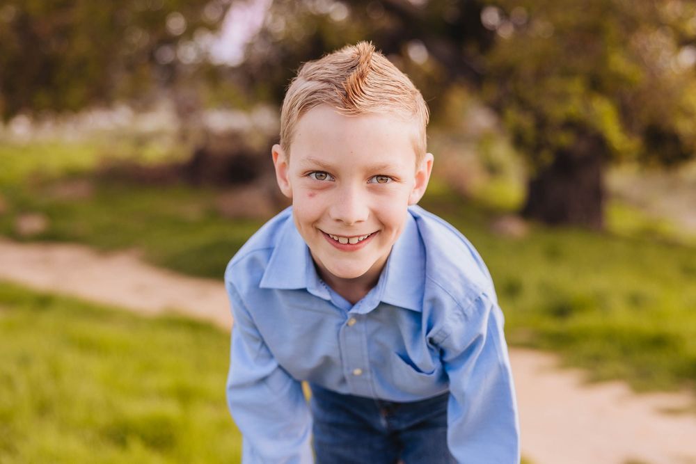 Boy in light blue polo shirt smiles brightly at camera against green grass background.