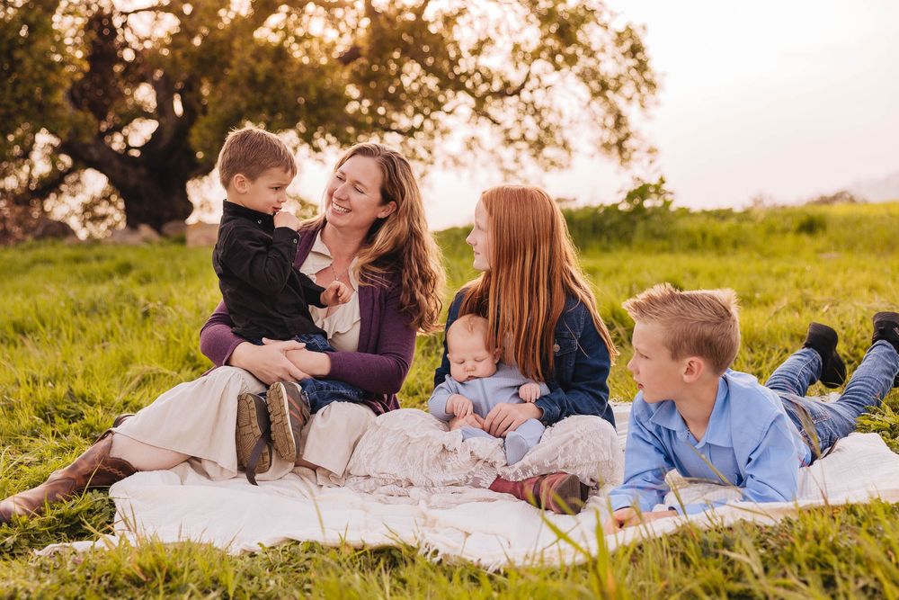 Family of five relaxing together on white blanket in grassy field at sunset.