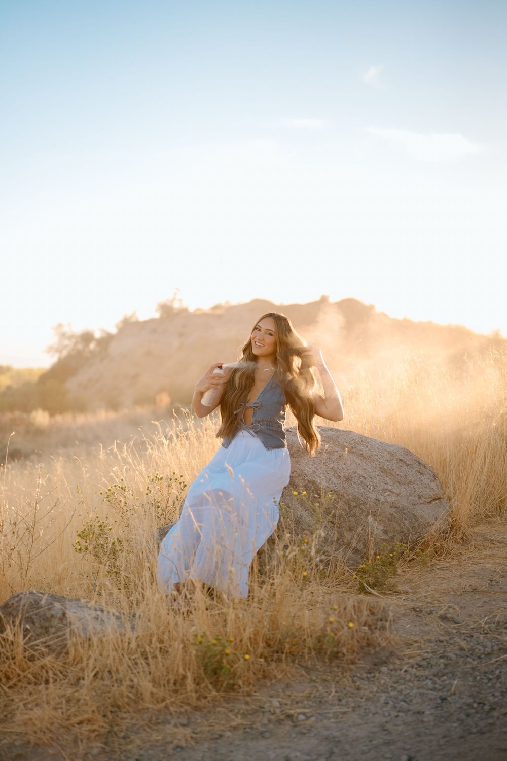 Woman in flowing white dress sits on large boulder at golden sunset in dry grassy landscape.